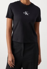 Sort bomulds T-shirt med rund hals, korte ærmer og et centralt hvidt logo, der viser "CK Jeans." Beskåret design med en glat tekstur.