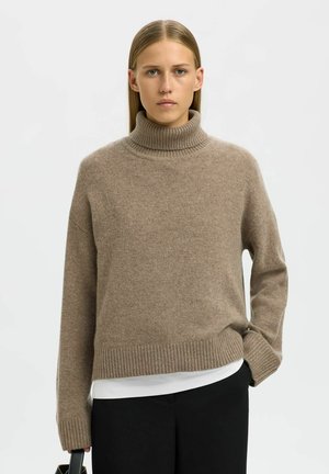 Pull à col roulé beige en tricot doux avec un col et un ourlet côtelés. Manches larges avec une coupe décontractée, associé à un t-shirt blanc et un pantalon noir.