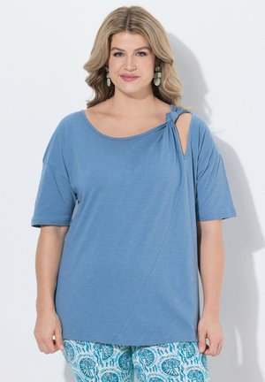 Frau mit welligem blondem Haar, die ein hellblaues Kurzarmoberteil mit Knotendetail an der Schulter und blaue gemusterte Hose trägt und vor weißem Hintergrund steht.