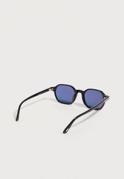 Slnečné okuliare s čiernym rámom a geometrickými modrými sklami ležia otvorené na bielom povrchu.