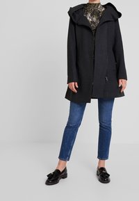 Cappotto nero con cappuccio, maniche lunghe e chiusura a cerniera. Presenta una vestibilità rilassata e un tessuto strutturato. Abbinato a jeans blu e scarpe nere.