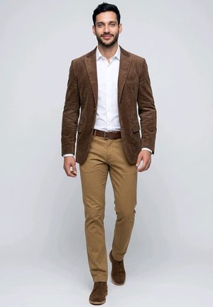 Hombre caminando con una chaqueta de pana marrón, camisa blanca, pantalones beige y zapatos de ante marrones sobre un fondo liso gris claro.