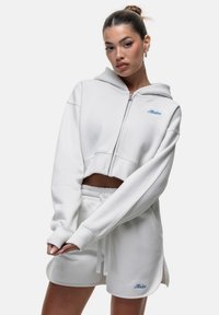 Felpa bianca croppata con zip e tasca frontale, abbinata a pantaloni corti bianchi con coulisse. Presenta un branding sottile in blu e oro. Tessuto morbido.