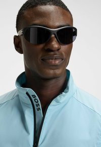 Sunglasses with black lenses and gray frames. Worn with a light blue jacket featuring a high collar and zip, labeled "BOSS" on the front.