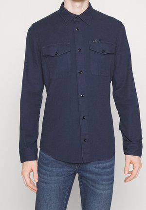 Chemise à manches longues de couleur navy avec fermeture à boutons, deux poches poitrine et un col classique. Tissu lisse avec une légère texture. Boutons noirs.