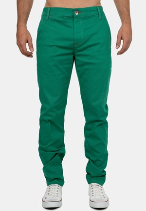 Chinos en coton verts à coupe slim, avec des accents de couture rouge et une fermeture à bouton. Associés à des baskets blanches.