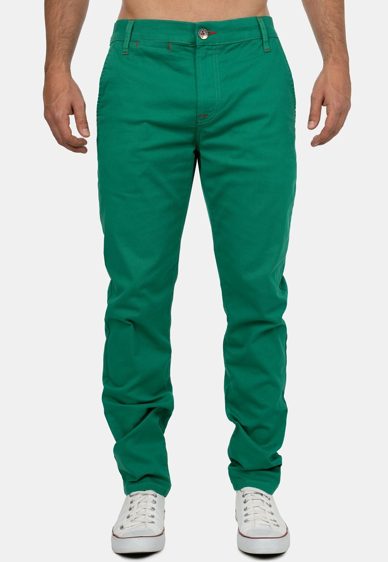 Chino in cotone verde con vestibilità slim, caratterizzati da dettagli di cucitura rossa e chiusura con bottone. Abbinati a sneakers bianche.
