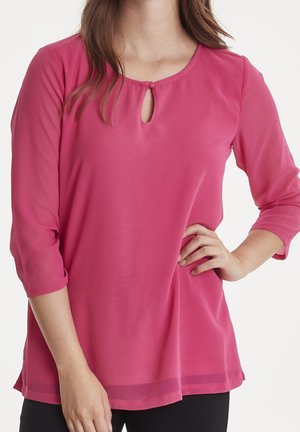 Vrouw met een roze blouse met lange mouwen, voorzien van een klein sleutelgat en knoopdetail bij de halslijn, poserend met één hand op de heup.
