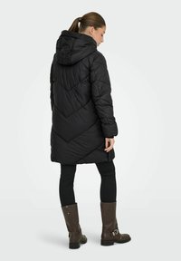 Cappotto imbottito trapuntato nero con cappuccio, caratterizzato da cuciture a zigzag, design lungo fino ai fianchi e tasche laterali. Indossato con leggings neri e stivali marroni.