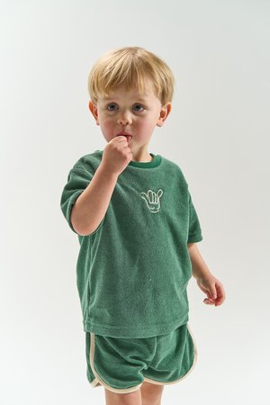 Kleiner Junge in grünem T-Shirt und Shorts isst einen kleinen Snack und steht vor einem schlichten hellen Hintergrund.