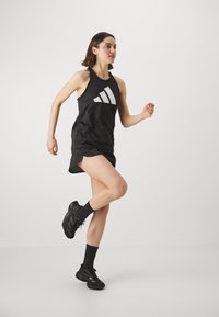 Černý sportovní tílko s bílým logem, spojené s černými šortkami, černými botami se zelenými akcenty a černými ponožkami. Osoba v pohybu.