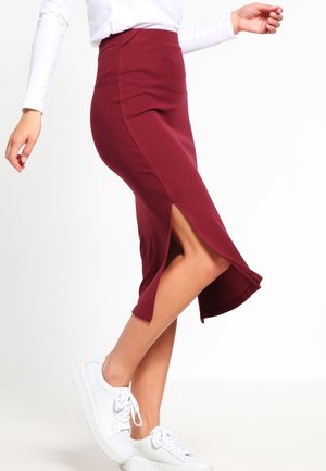 Kobieta w dopasowanej bordowej spódnicy midi z bocznym rozcięciem, białej bluzce z długim rękawem i białych trampkach, stojąca na jednolitym tle.