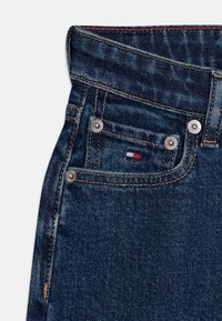 Vaqueros de mezclilla azul oscuro con un diseño ajustado. Las características incluyen herrajes plateados, un pequeño logotipo en rojo, blanco y azul, y costuras en contraste.