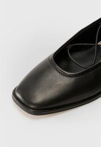 Shoe preto de couro plano com design de bico quadrado, apresentando uma tira fina no tornozelo e textura suave. Construção simples e elegante.