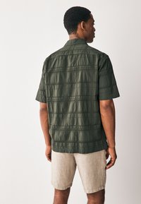 Dunkelgrünes Kurzarmhemd mit strukturiertem Muster, von hinten gezeigt, kombiniert mit hellbeigen Shorts. Einfacher Kragen und lockere Passform.