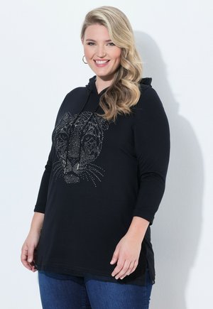 Lächelnde Frau mit langen blonden Haaren trägt schwarzen Hoodie mit Katzenmotiv aus Strasssteinen, blaue Jeans und Creolen vor weißem Hintergrund.