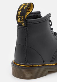 Bottine en cuir noir avec des coutures jaunes le long de la semelle, finition texturée et une languette jaune avec le texte "Bouncing Soles".