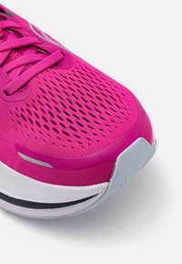 Chaussure de sport rose avec une tige en mesh respirant, surface texturée et accents réfléchissants. Présente une semelle blanche et grise avec des rayures noires.