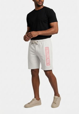 Uomo in piedi che indossa una t-shirt nera, pantaloncini bianchi con coulisse e la scritta rosa "CHABRAND" sul lato, e sneakers beige.