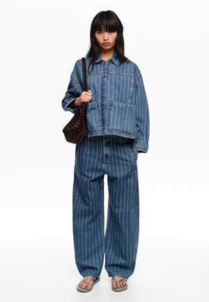 Joven mujer con chaqueta y pantalones de mezclilla azul con rayas finas, llevando un bolso de hombro oscuro tejido y sandalias grises.