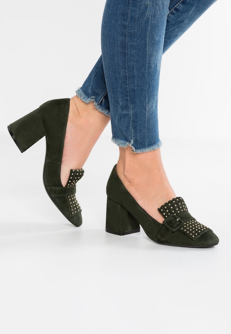 Pieds portant des chaussures à talons blocs en daim vert foncé avec un nœud clouté et un détail de boucle, associées à un jean en denim bleu à ourlet effiloché.