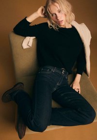 Tmavomodré riflové nohavice so svetlým prešitím v kombinácii s čiernym svetrom. Modelka sedí na zelenej stoličke na hnedom pozadí.