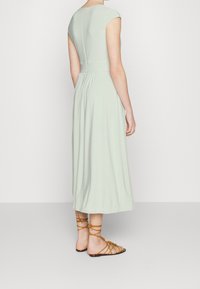Světlé zelené midi šaty s krátkým rukávem, uzkým pasem a na zadní straně s řasenou texturou; kombinováno s hnědými sandály na pásku.
