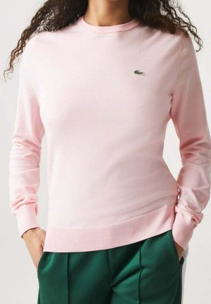 Femme portant un pull à manches longues rose clair avec un petit logo sur la poitrine et un pantalon vert foncé, les mains dans les poches, cheveux bouclés visibles.