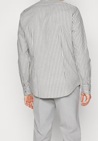 Overhemd met lange mouwen met dunne verticale zwarte en witte strepen, een afgeronde zoom en manchetten met knopen, gecombineerd met lichtgrijze broek.