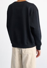 Sudadera azul oscuro con cuello redondo, corte relajado, puños y dobladillo acanalados. Tejido suave con un mínimo de logotipos, combinada con pantalones beige claros.