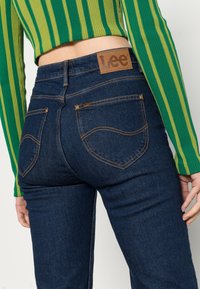 Jeans in denim blu scuro con cuciture arancioni e due tasche posteriori. Presenta un'etichetta in pelle marrone con il logo Lee sulla vita.