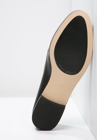 Chaussure en cuir noir avec une tige lisse et une semelle en caoutchouc plate. Comprend un emblème de marque et un motif de relief texturé sous la semelle.