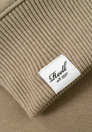 Beige ribbed knit fabric with a white rectangular label stitched on, displaying the brand name "Reell" and "est. 1997" in black script.