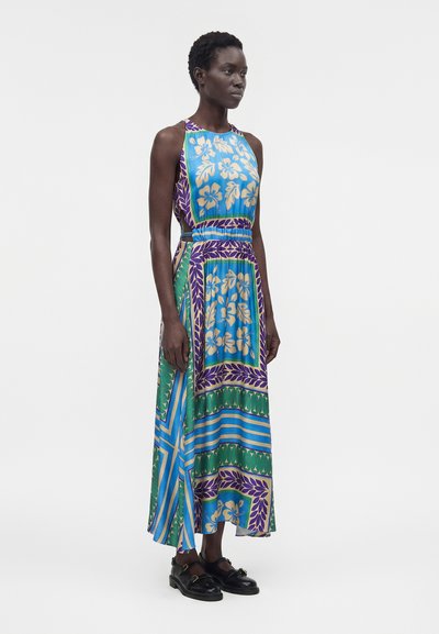 Γυναίκα φοράει αμάνικο, μακρύ, αέρινο φόρεμα με μπλε, πράσινα, μωβ και μπεζ λουλουδάτα και γεωμετρικά σχέδια, συνδυασμένο με μαύρα loafers.