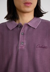 Polo majica prugasta ljubičasta boja, s ovratnikom, tri gumba i izvezenim "Carhartt" logotipom. Napravljena je od mekog materijala s suptilnom teksturom.