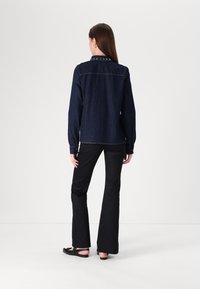 Camicia di jeans blu scuro con maniche lunghe, polsini con bottoni e borchie decorative lungo il colletto. Indossata con pantaloni neri a zampa e sandali bassi.