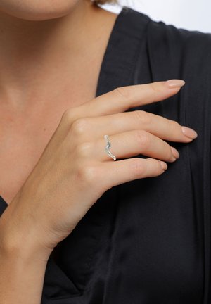Sølv bølgete ring båret på en finger, med en glat overflade og subtile detaljer, mod en mørk stofbaggrund.