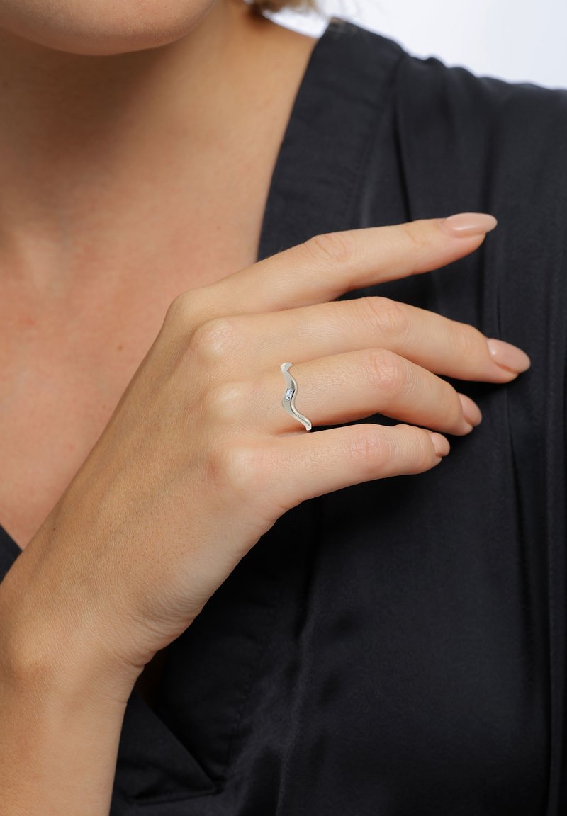 Zilveren golfvormige ring gedragen om een vinger, met een gladde textuur en subtiele details, tegen een donkere stof achtergrond.