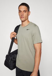 Olive rohelise värvi Nike t-särk, ümmarguse kaelusega, lühikeste varrukatega ja musta logoga; mudel hoiab käes musta spordikotti, millel on tekstuuriga detailid.