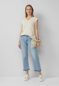 Vrouw staat tegen een muur en draagt een crèmekleurig V-hals T-shirt, lichtblauwe cropped jeans, witte sneakers en houdt een beige geweven handtas vast.