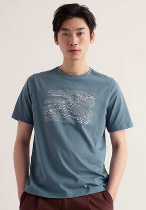 Mann steht frontal, trägt ein blaues T-Shirt mit weißem abstraktem Liniendesign und braune Hose, vor einem schlichten hellen Hintergrund.