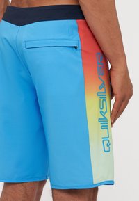 Kék úszónadrág, amelyen átmenetes mintázat látható narancssárga és sárga kiegészítőkkel. Zsebet és Quiksilver logót tartalmaz az oldalon.
