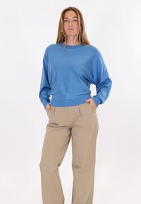 Žena stojící s rukama v kapsách, oblečená v modrém topu s dlouhým rukávem a béžových kalhotách s širokými nohavicemi na bílém pozadí.