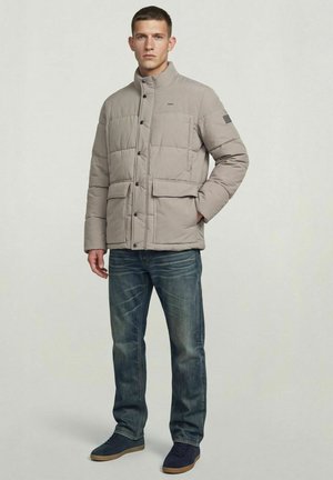 Hombre con chaqueta acolchada beige con bolsillos con solapa, vaqueros azules y zapatillas náuticas, de pie con una mano en el bolsillo.