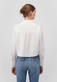 White long-sleeve shirt featuring a textured fabric, back yoke seam, and buttoned cuffs, paired with high-waisted blue jeans.