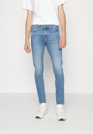 Mann trägt hellblaue Slim-Fit-Jeans, weißes T-Shirt, weiße Socken und weiße Sneaker und steht vor einem einfarbigen hellen Hintergrund.