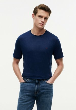Junger Mann mit kurzen braunen Haaren, der ein marineblaues T-Shirt und blaue Jeans trägt, steht mit den Händen in den Taschen vor einem einfarbigen Hintergrund.