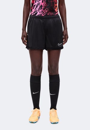 Άτομο που φοράει μαύρο σορτς ποδοσφαίρου Nike, μαύρες κάλτσες Nike μέχρι το γόνατο, πορτοκαλί ποδοσφαιρικά παπούτσια και ροζ και μαύρο πουκάμισο με σχέδια.