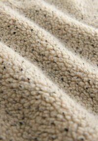 Beige fleecestof met een gestructureerd oppervlak, voorzien van een zachte, pluizige uitstraling en verspreide zwarte spikkels.