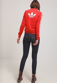 Červená sportovní bunda s bílými pruhy a logem na zadní straně, kombinovaná s tmavými džíny a hnědými šněrovacími botami.